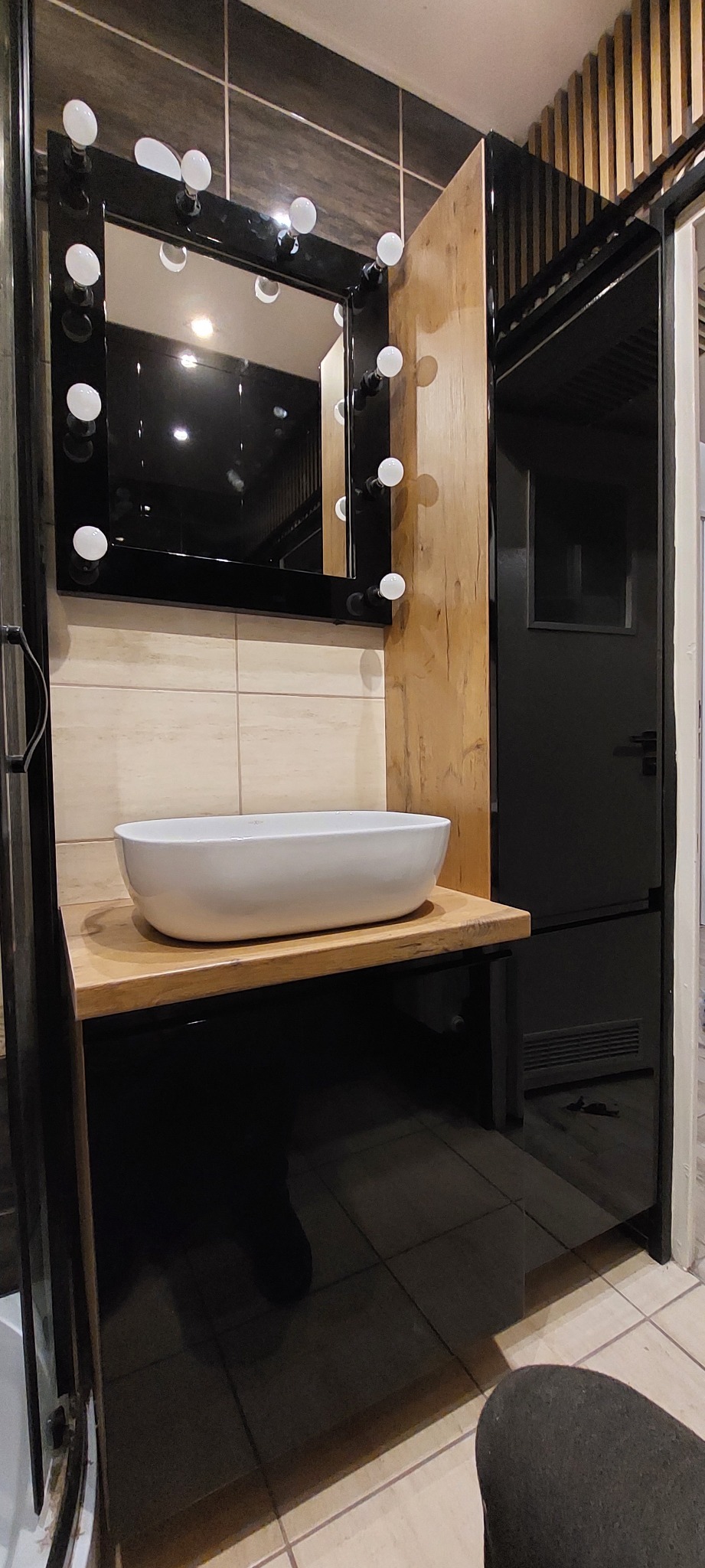 Nowoczesna łazienka z umywalką nablatową, lustrem podświetlanym żarówkami, drewnianym blatem i czarnymi szafkami na wysoki połysk.