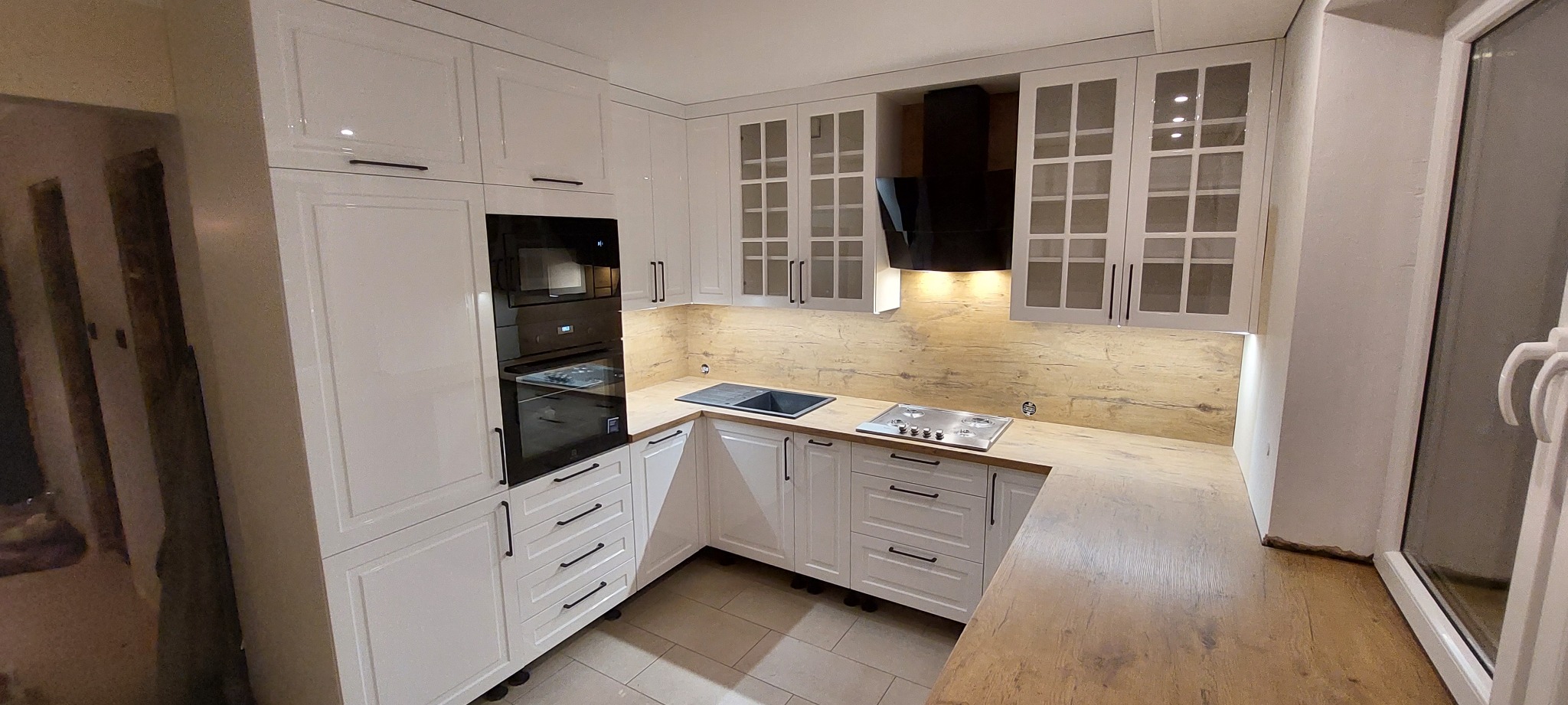 Nowoczesna, jasna kuchnia z białymi, lakierowanymi frontami, czarnymi uchwytami, drewnianym blatem i zabudowanym piekarnikiem oraz płytą gazową.