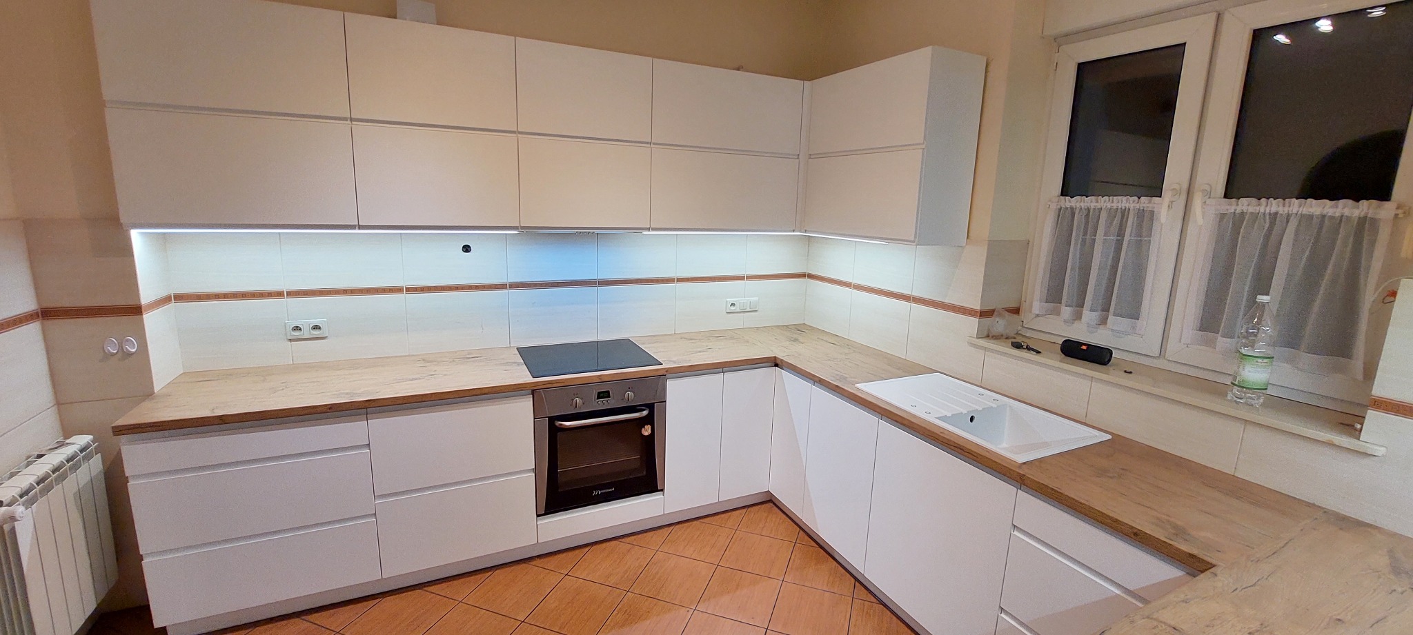 Nowoczesna kuchnia w kształcie litery L z białymi frontami szafek i blatem imitującym drewno, oświetlona listwą LED pod szafkami wiszącymi, z płytą indukcyjną i piekarnikiem.