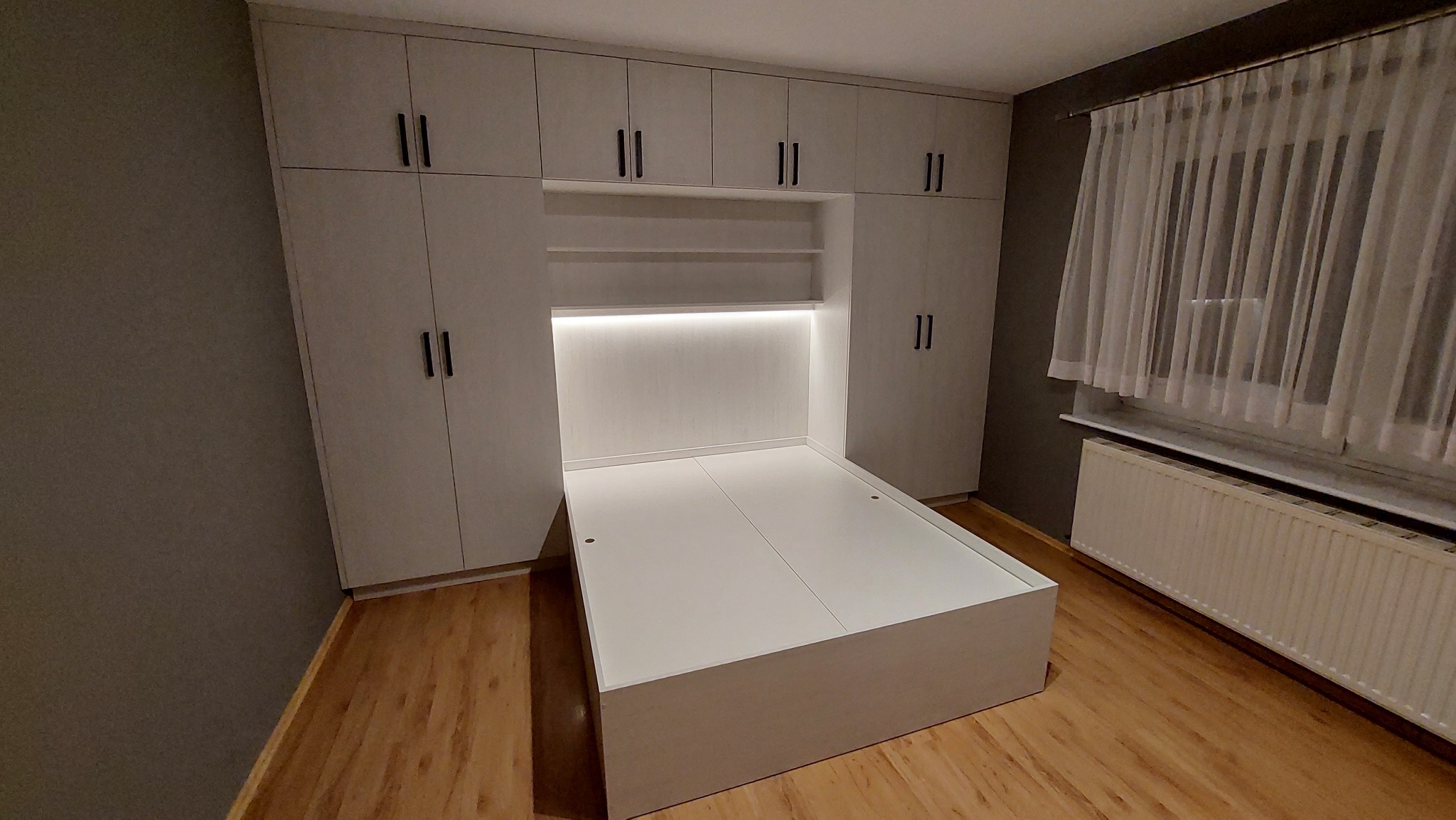 Biała zabudowa meblowa z szafami i wnęką z podświetleniem LED, tworząca ramę dla łóżka, na podłodze panele drewnopodobne.