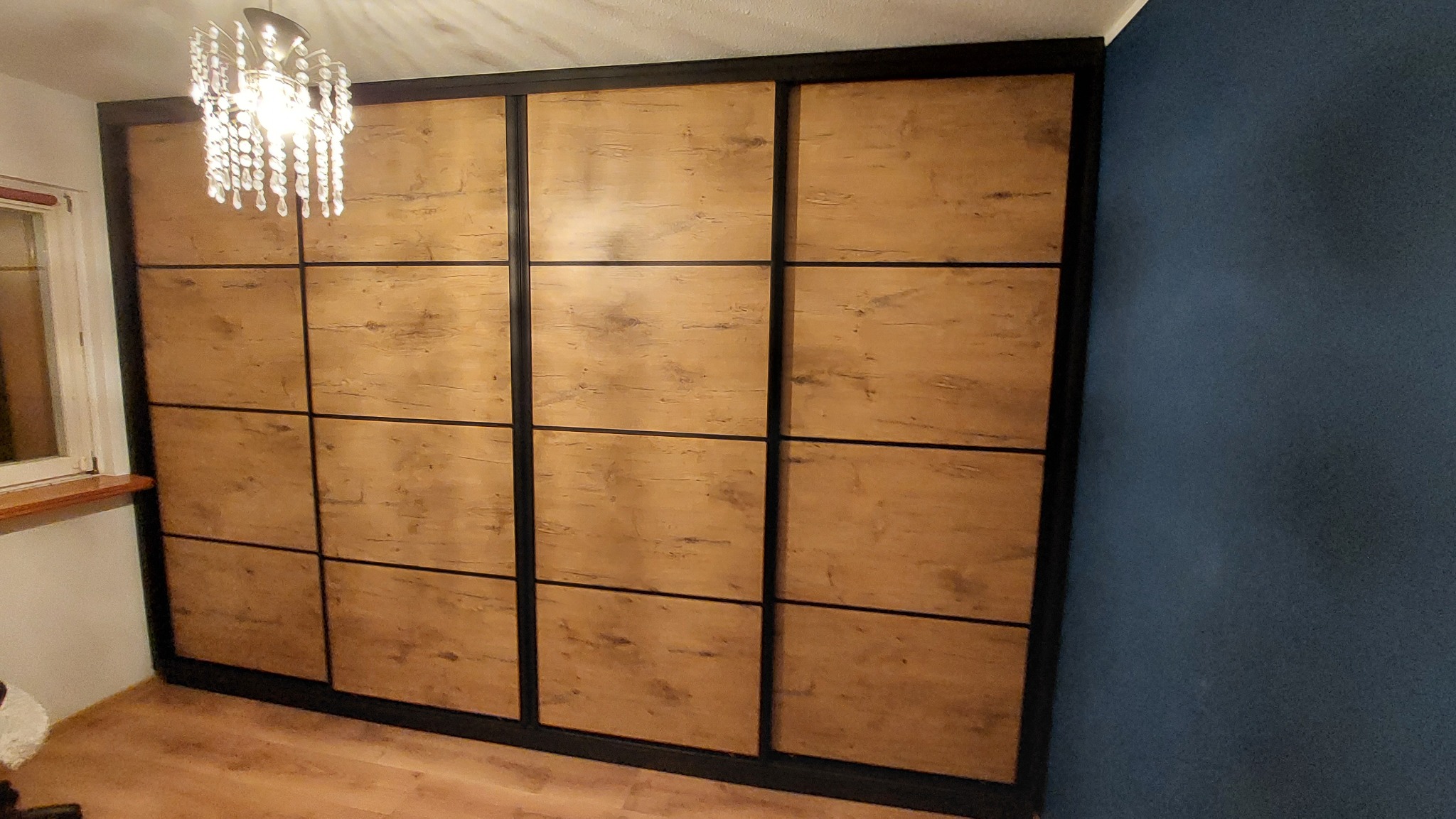 Duża szafa wnękowa z przesuwnymi drzwiami o drewnianej fakturze, podzielona na kwadratowe panele z czarnymi ramami, oświetlona przez kryształowy żyrandol.