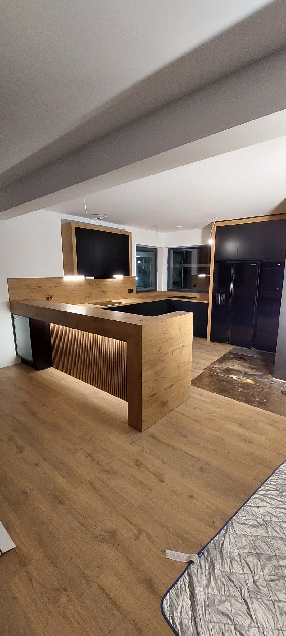 Nowoczesna kuchnia z wyspą o drewnianym wykończeniu i oświetleniem LED, czarne szafki w tle, podłoga laminowana, widoczne elementy wykończeniowe.