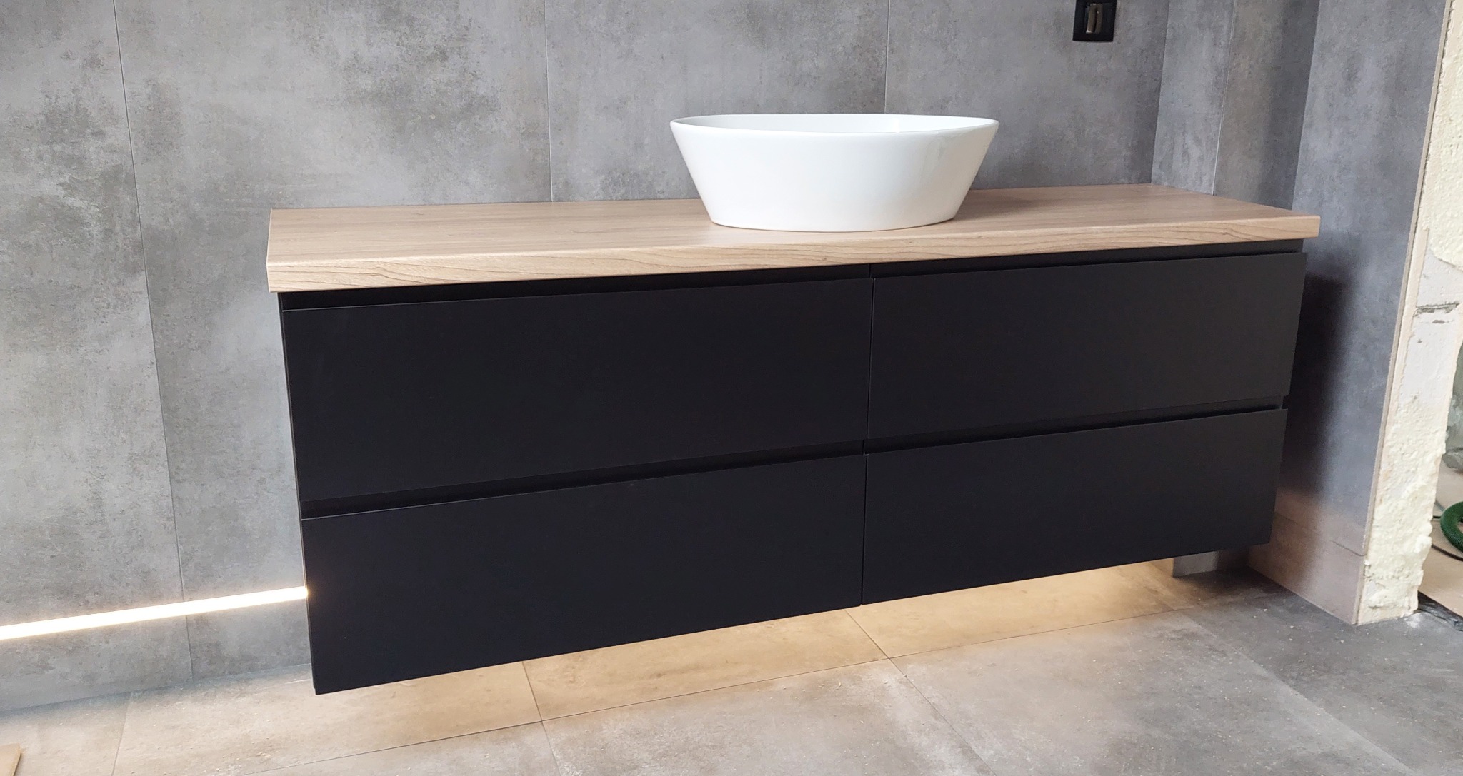 Czarna szafka łazienkowa z drewnianym blatem i białą umywalką nablatową, oświetlenie LED pod szafką, szare płytki na ścianie i podłodze, widoczny fragment niezakończonego wykończenia ściany.