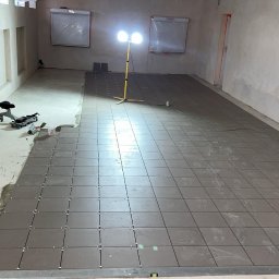 Bouwtornado - Układanie szarych płytek podłogowych w dużym pomieszczeniu z surowymi ścianami. Widoczne narzędzia glazurnicze i lampa oświetlająca obszar roboczy.