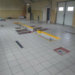 Bouwtornado - Wyłożona jasnymi płytkami podłoga warsztatu z kanałami naprawczymi, zabezpieczonymi żółtymi krawędziami. Widoczne kratki odpływowe i elementy wyposażenia warsztatu w tle.