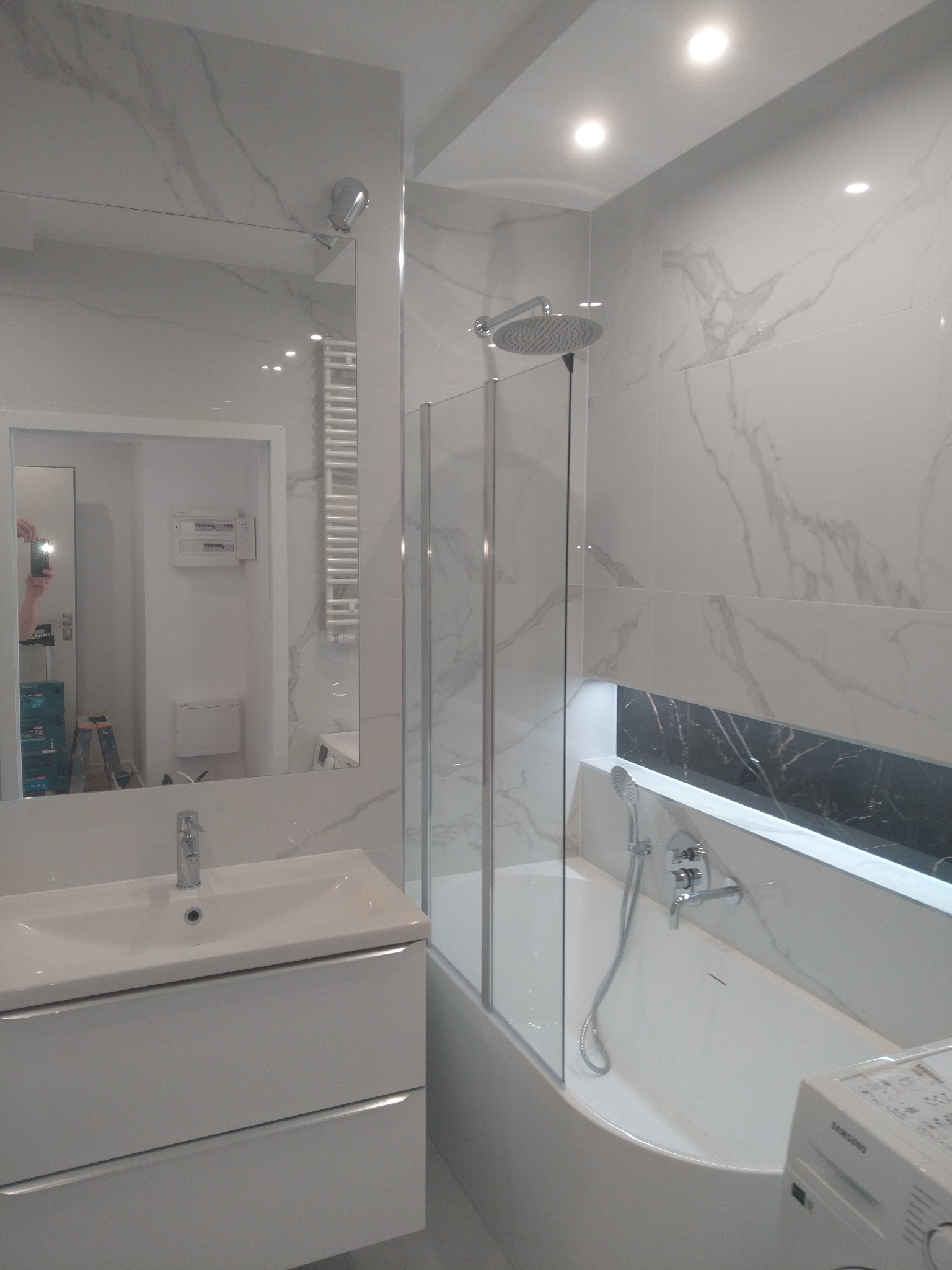 Nowoczesna łazienka z białymi marmurkowymi płytkami, kabiną prysznicową z szybą, umywalką z szafką i wanną z czarnym akcentem wnęki, widoczne odbicie osoby robiącej zdjęcie w lustrze.