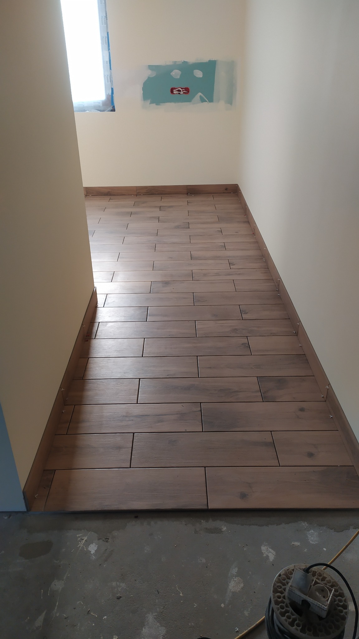 Ułożone brązowe płytki podłogowe imitujące drewno w korytarzu, widoczne krzyżyki dystansowe przy listwie przypodłogowej, ściany w kolorze złamanej bieli, fragment surowej podłogi z resztkami zaprawy.