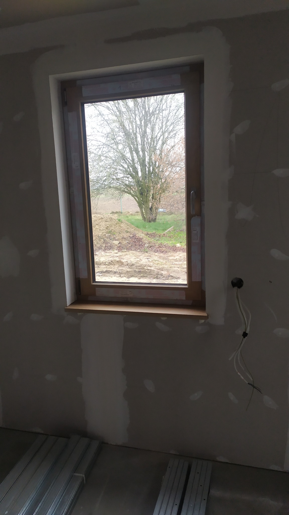 Ściana z oknem po nałożeniu gładzi, widoczne niedoskonałości i kable wystające ze ściany, profile metalowe na podłodze.