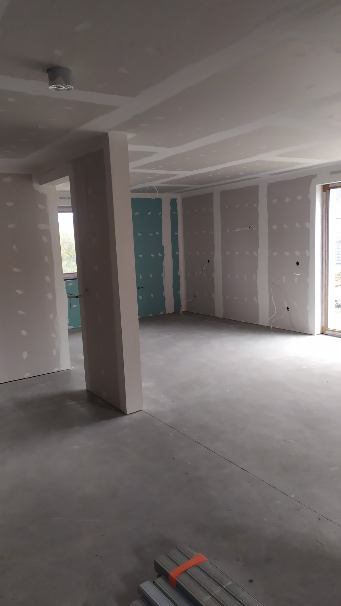 Surowe wnętrze z płytami kartonowo-gipsowymi na ścianach i suficie, widoczne łączenia płyt, podłoga betonowa, profile metalowe na podłodze w rogu, okno w tle.