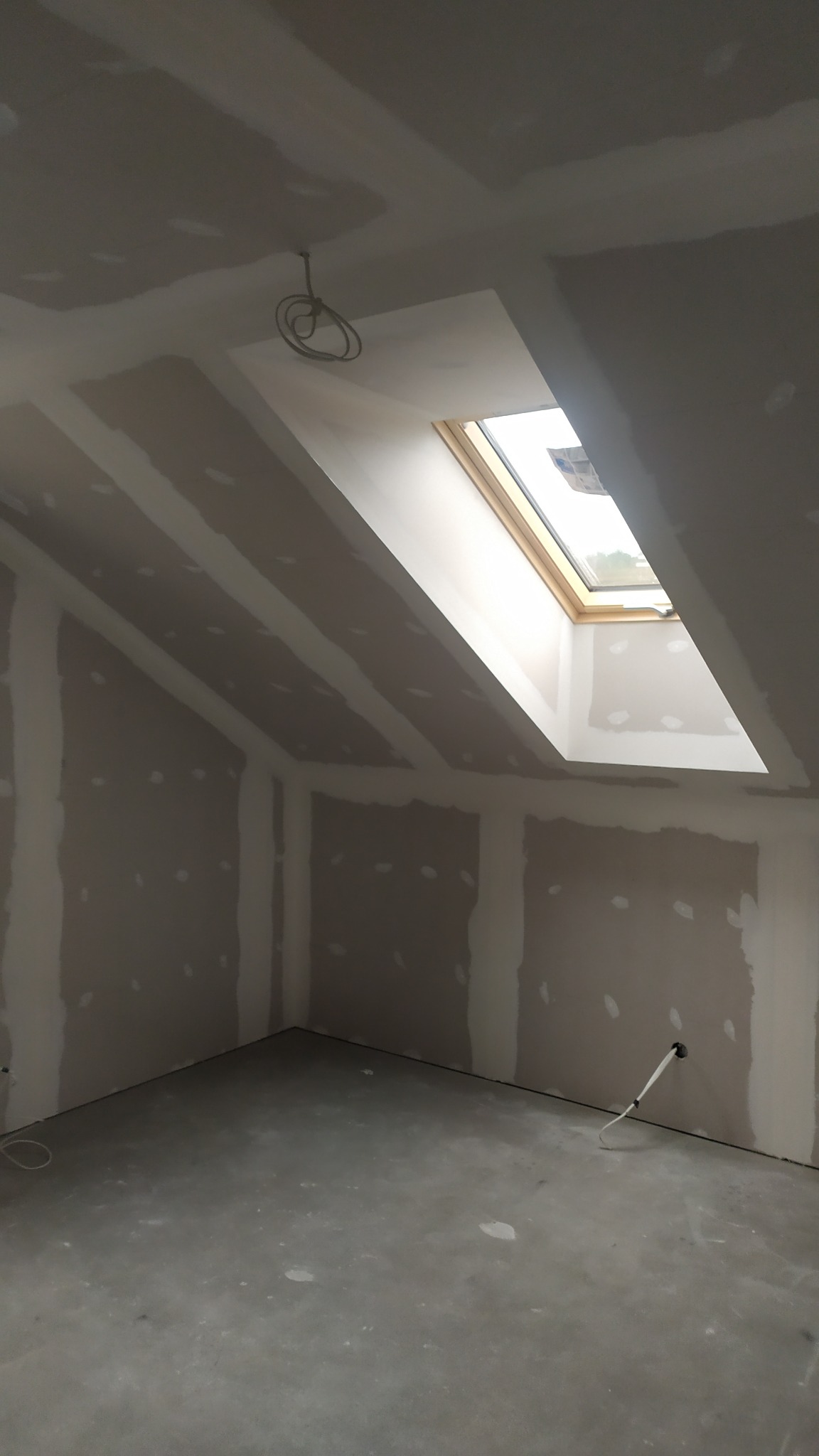 Poddasze po montażu płyt karton-gips z oknem dachowym i wystającymi przewodami elektrycznymi, przygotowane do dalszych prac wykończeniowych.