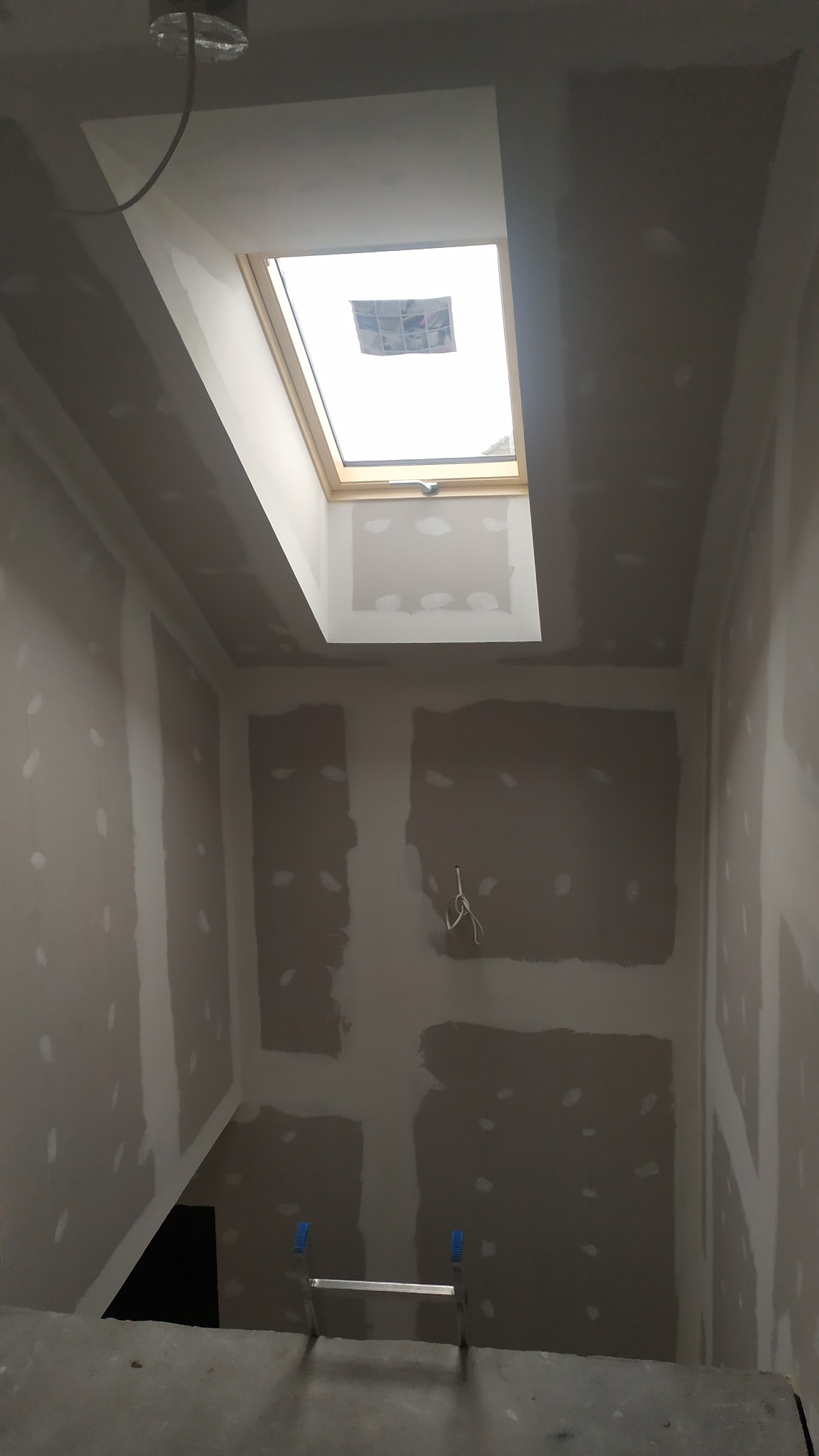 Wnętrze w trakcie prac, widoczne ściany z płyt kartonowo-gipsowych, częściowo zaszpachlowane, okno dachowe i drabina na podłodze.