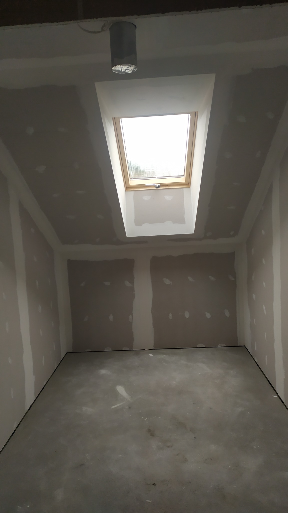 Pokój w trakcie prac wykończeniowych: ściany z płyt kartonowo-gipsowych z widocznymi łączeniami, sufit z oknem dachowym, surowa betonowa podłoga i prowizoryczne oświetlenie.