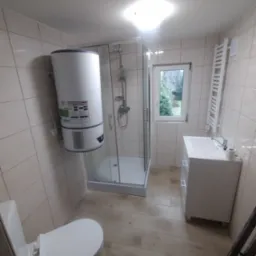 Nowoczesna łazienka z kabiną prysznicową, wiszącym bojlerem Ariston, grzejnikiem drabinkowym i białą umywalką z szafką, widziana z niskiego kąta, ukazująca fragment drabiny.