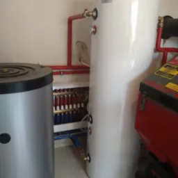 Instalacja kotła grzewczego z podłączeniem w Wielgomłynach, widoczne zbiorniki buforowe, rury w kolorze czerwonym i niebieskim oraz rozdzielacz ogrzewania podłogowego.