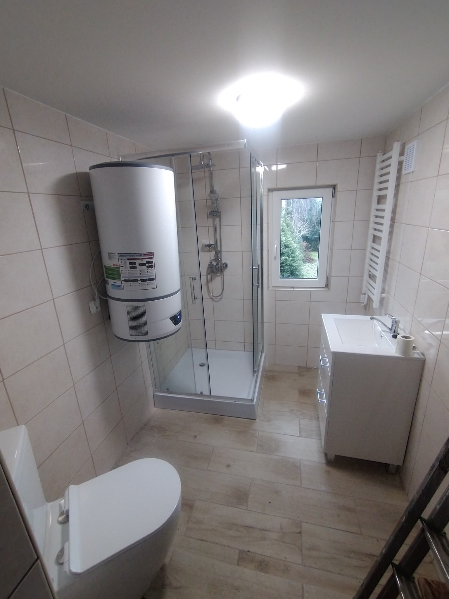 Nowoczesna łazienka z kabiną prysznicową, wiszącym bojlerem Ariston, grzejnikiem drabinkowym i białą umywalką z szafką, widziana z niskiego kąta, ukazująca fragment drabiny.