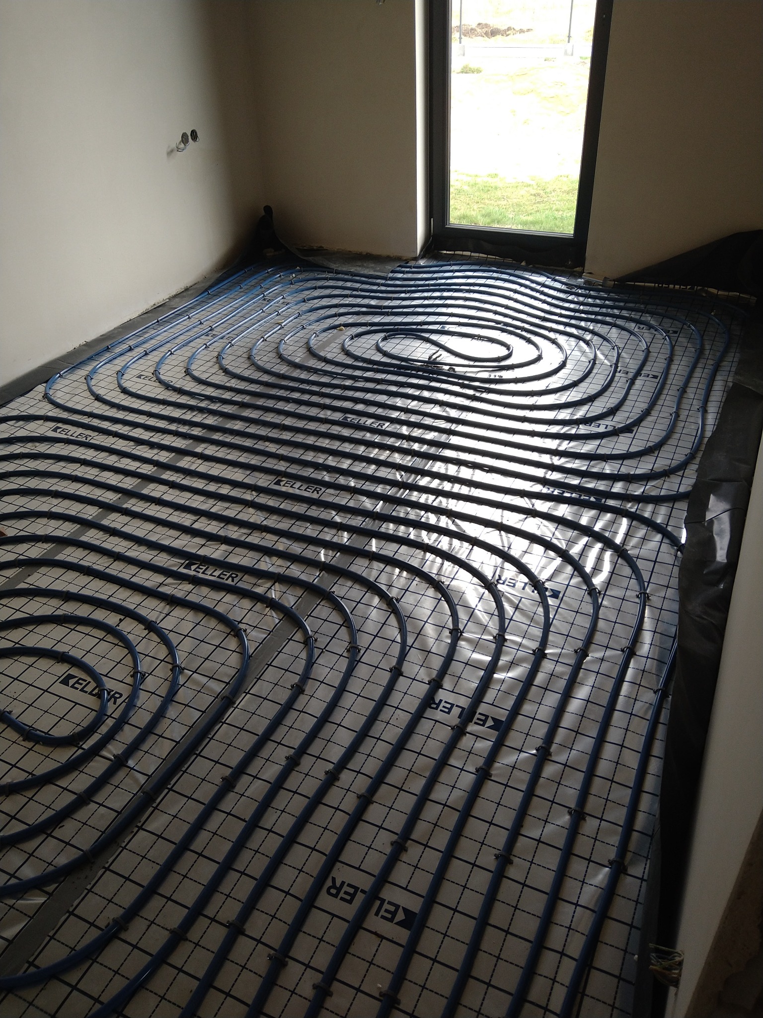 Niebieskie rury systemu ogrzewania podłogowego marki Keller ułożone spiralnie na folii z nadrukiem kratki w surowym wnętrzu z oknem.