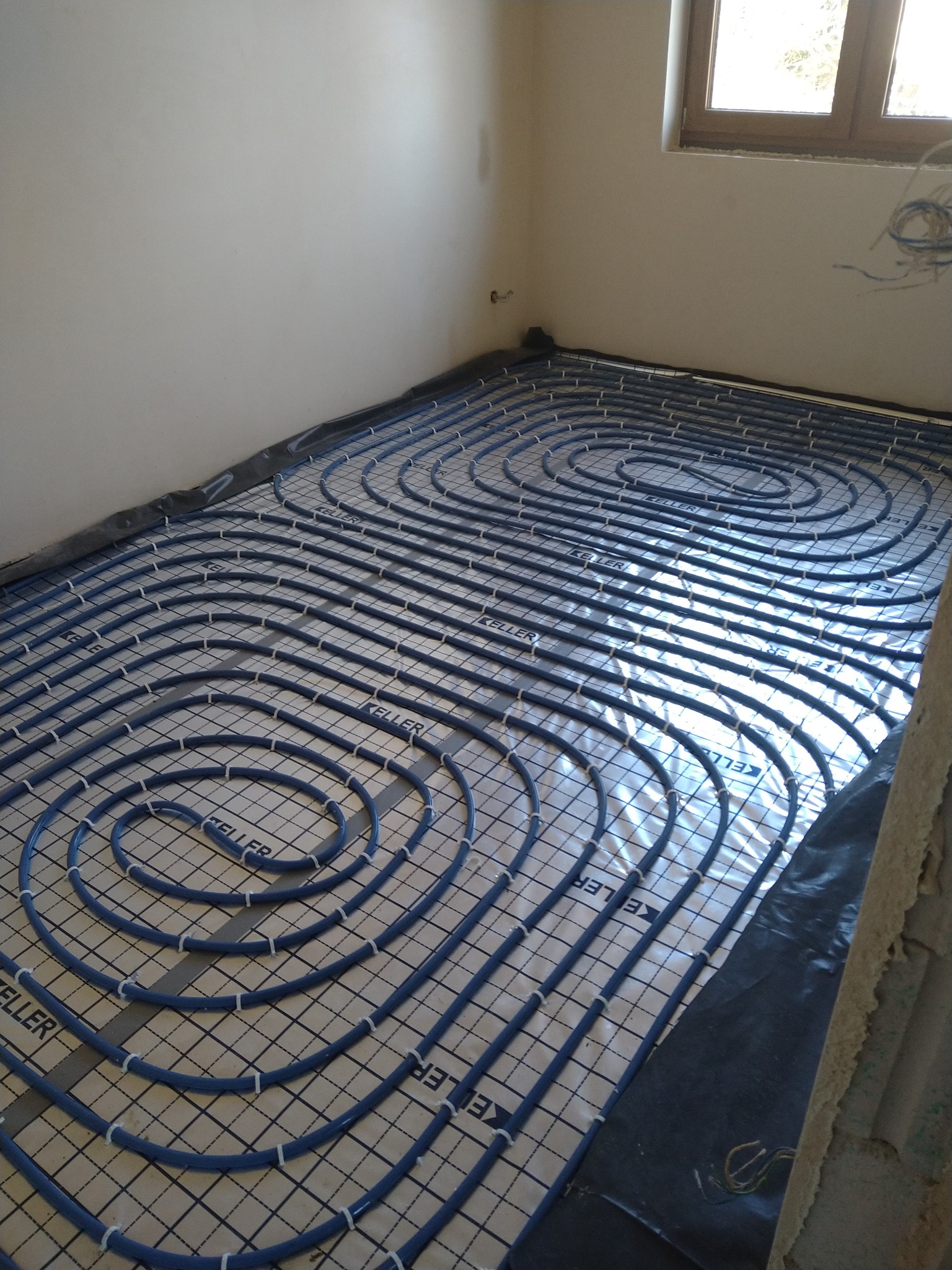 Niebieskie rury ogrzewania podłogowego marki Keller rozłożone na podłodze w nowym budownictwie, przymocowane białymi uchwytami do siatki montażowej. Widoczne okno i fragment ściany w surowym stanie.