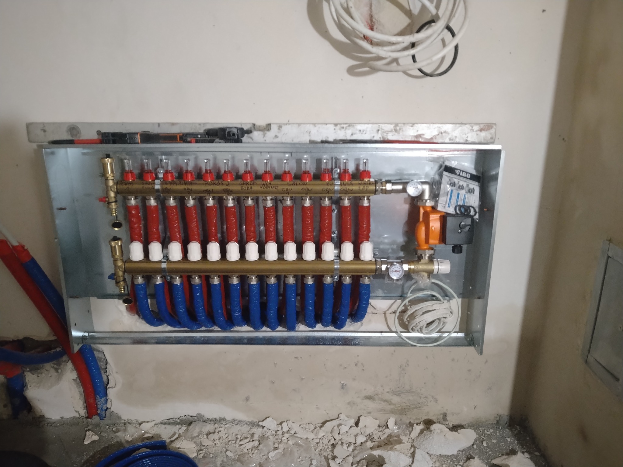 Rozdzielacz ogrzewania podłogowego w metalowej obudowie, z czerwonymi i niebieskimi rurami, zamontowany na surowej ścianie w trakcie instalacji, widoczne narzędzia i poziomica na górze obudowy.