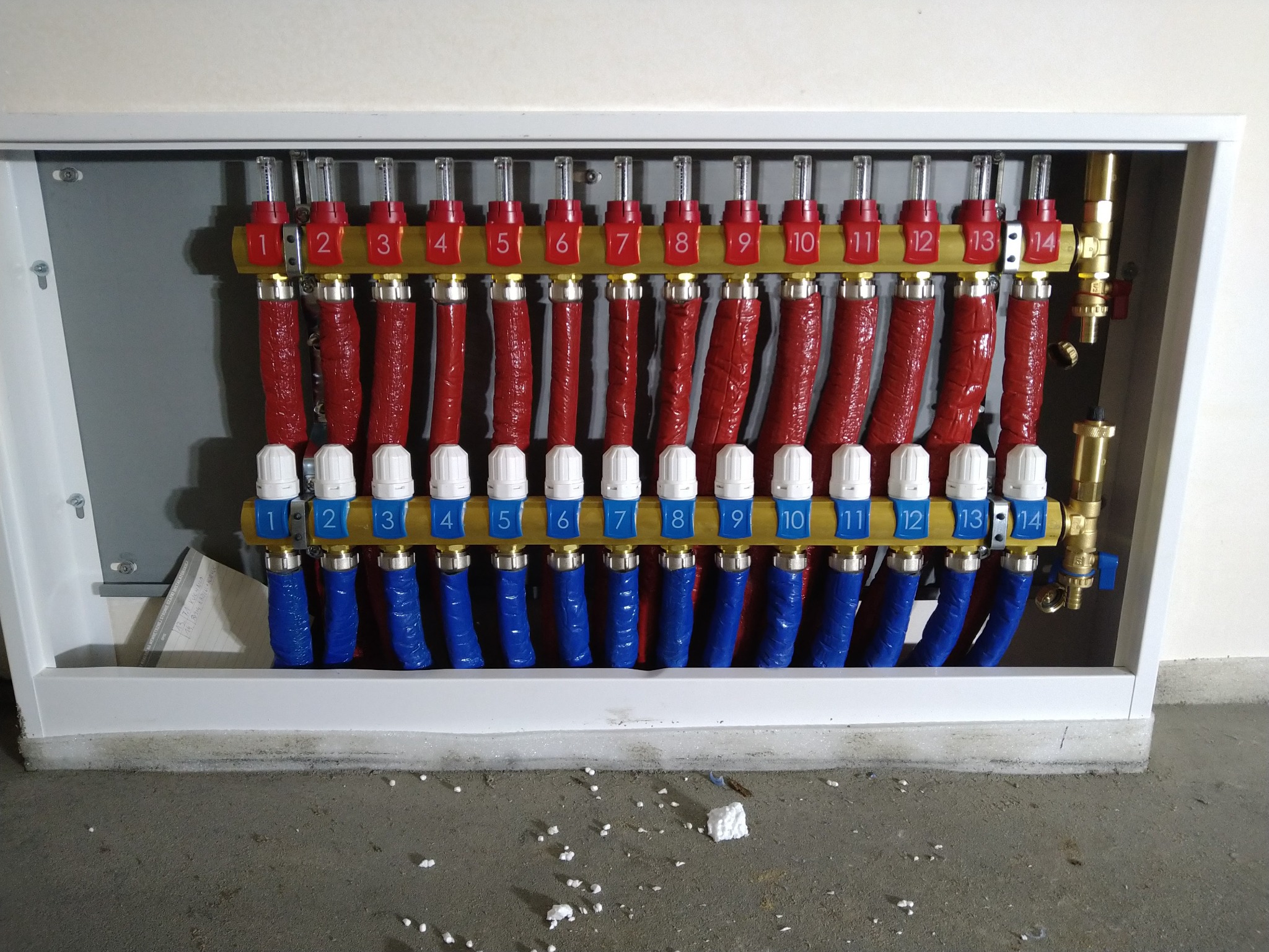 Rozdzielacz ogrzewania podłogowego z numerowanymi sekcjami w szafce instalacyjnej, rury doprowadzające i odprowadzające ciepło w izolacji, widoczne oznaczenia numerów obiegów grzewczych.