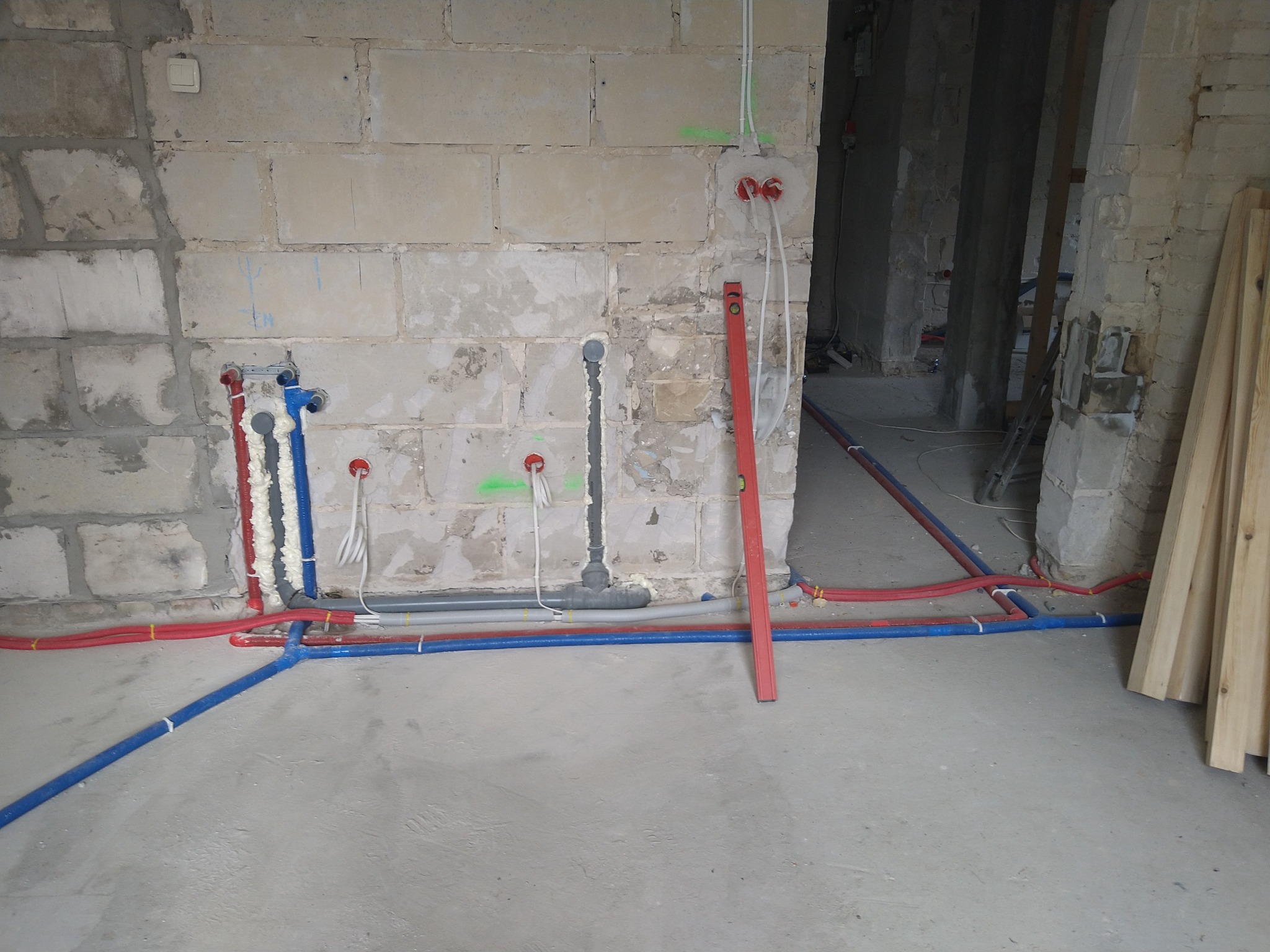Surowe wnętrze w trakcie prac instalacyjnych: widoczne kolorowe rury wodne i kanalizacyjne mocowane do ściany z betonu komórkowego, częściowo zabezpieczone pianką montażową, z poziomnicą opartą...