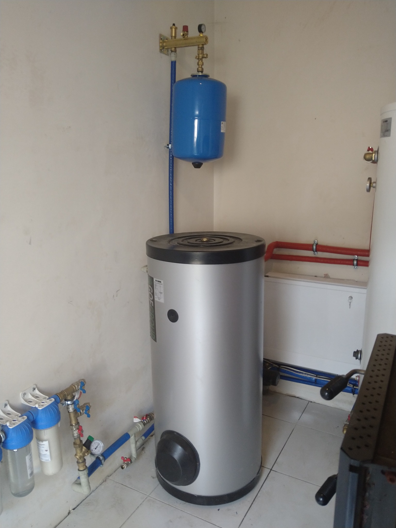 Szary bojler z niebieskim zbiornikiem wyrównawczym i filtrami wody w pomieszczeniu z białymi ścianami i płytkami podłogowymi.