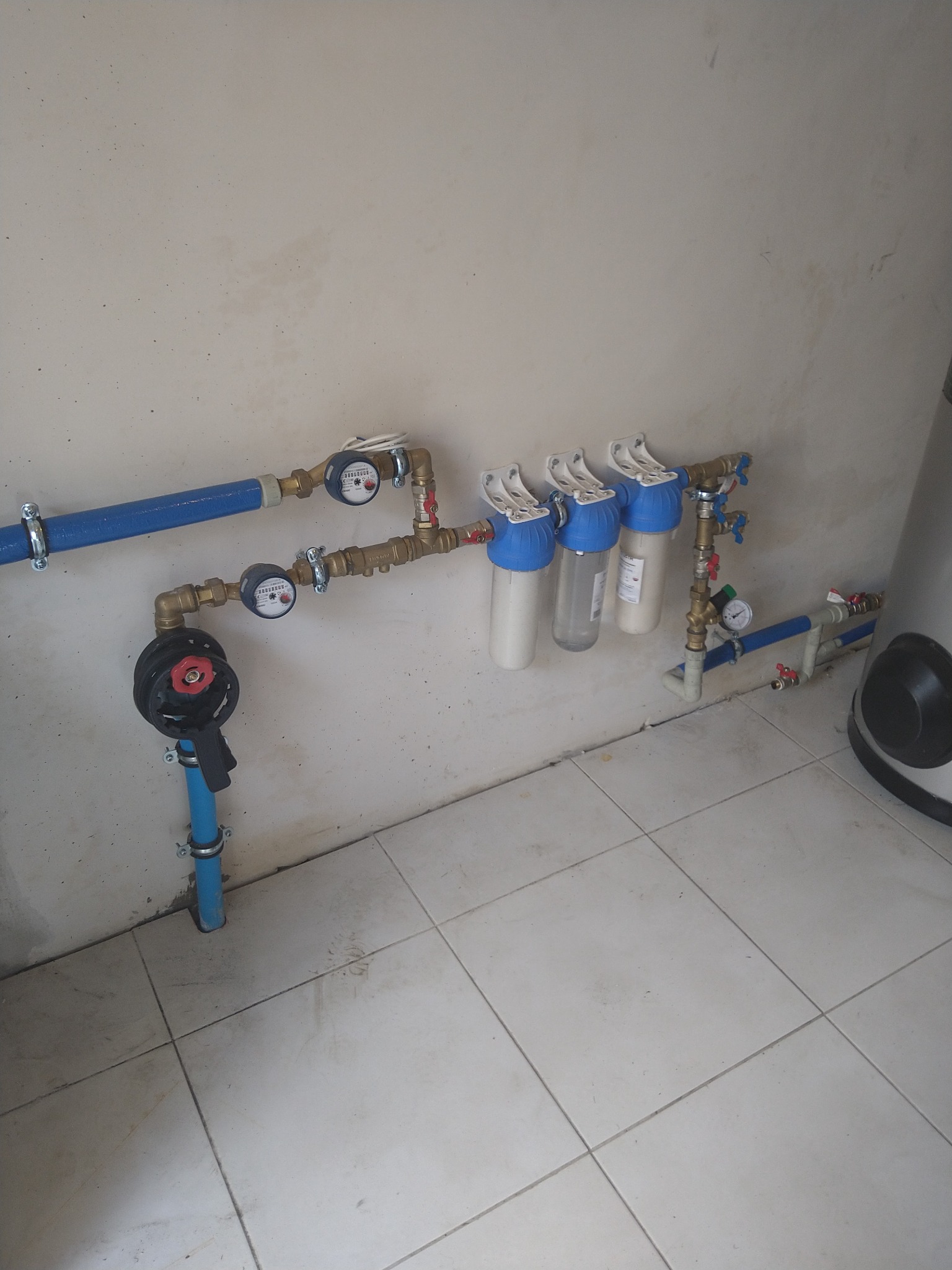 Zestaw filtrów wody, liczniki i zawór zamontowane na ścianie w pomieszczeniu z wyłożoną kafelkami podłogą, widoczne elementy instalacji wodnej.
