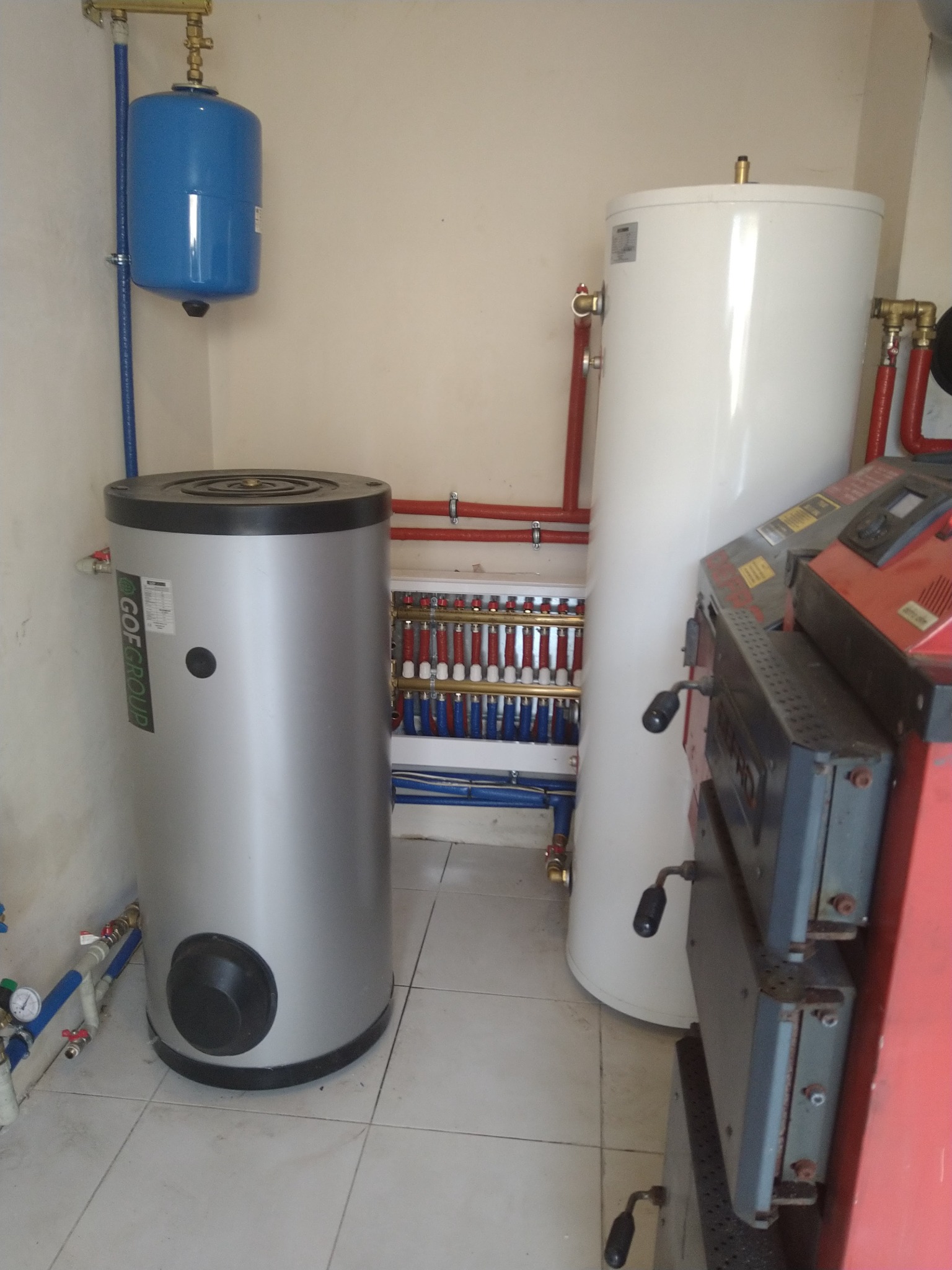 Kompaktowa kotłownia z dwoma zasobnikami ciepłej wody: srebrnym z czarnym akcentem i białym, obok rozdzielacza grzewczego z licznymi zaworami i rurami w kolorach czerwonym i niebieskim oraz pieca...