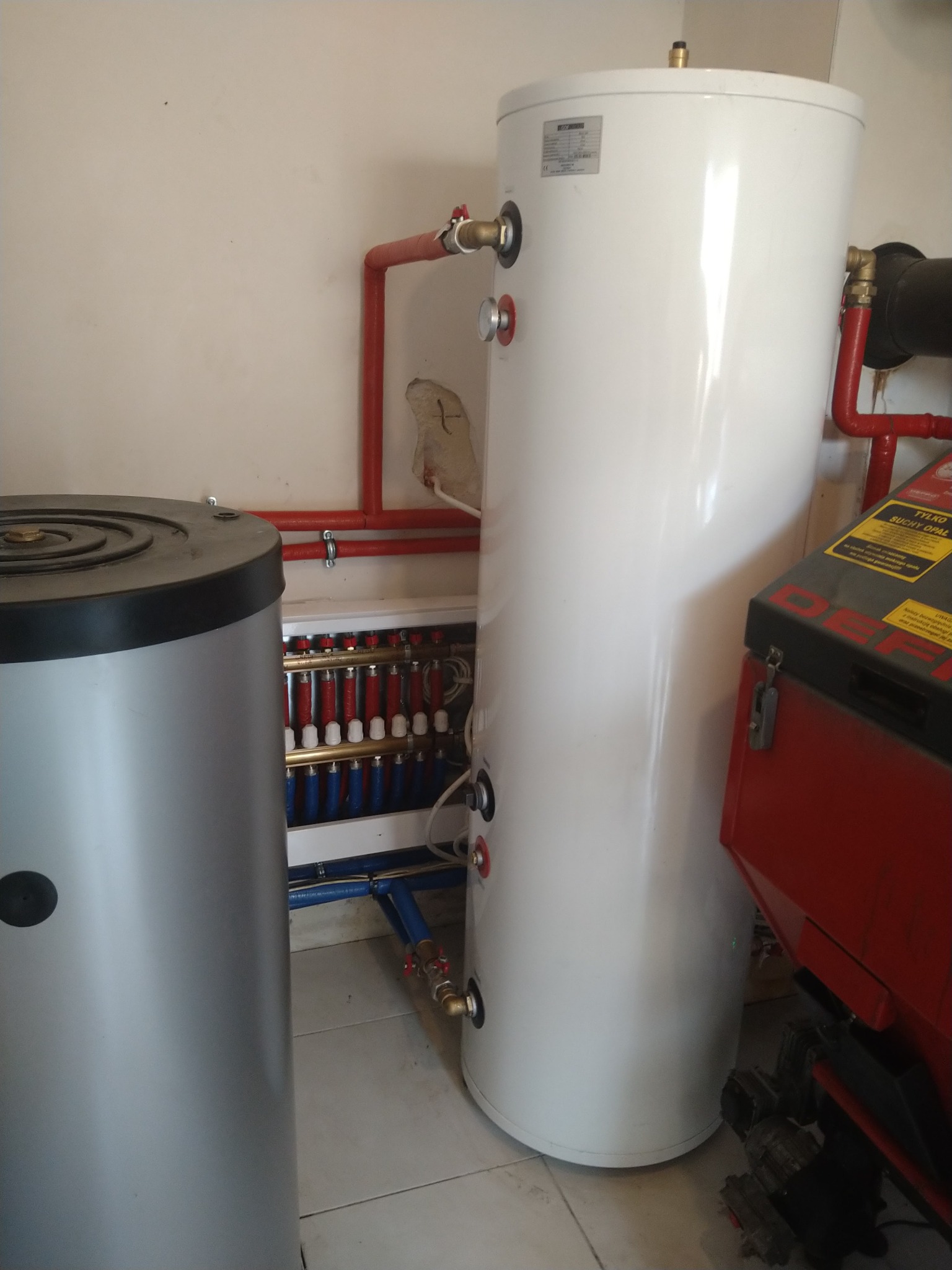 Instalacja kotła grzewczego z podłączeniem w Wielgomłynach, widoczne zbiorniki buforowe, rury w kolorze czerwonym i niebieskim oraz rozdzielacz ogrzewania podłogowego.