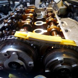 Setup Motors Serwis Samochodowy - Dobry Mechanik - Silnik samochodowy z widocznymi wałkami rozrządu, łańcuchem rozrządu i żółtym ślizgiem, podczas naprawy lub przeglądu, oświetlenie warsztatowe.