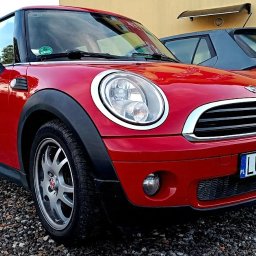 Setup Motors Serwis Samochodowy - Dobry Mechanik - Czerwony Mini Cooper zaparkowany na żwirowym parkingu przed warsztatem samochodowym, widoczny plakat Motul i Skoda w tle.