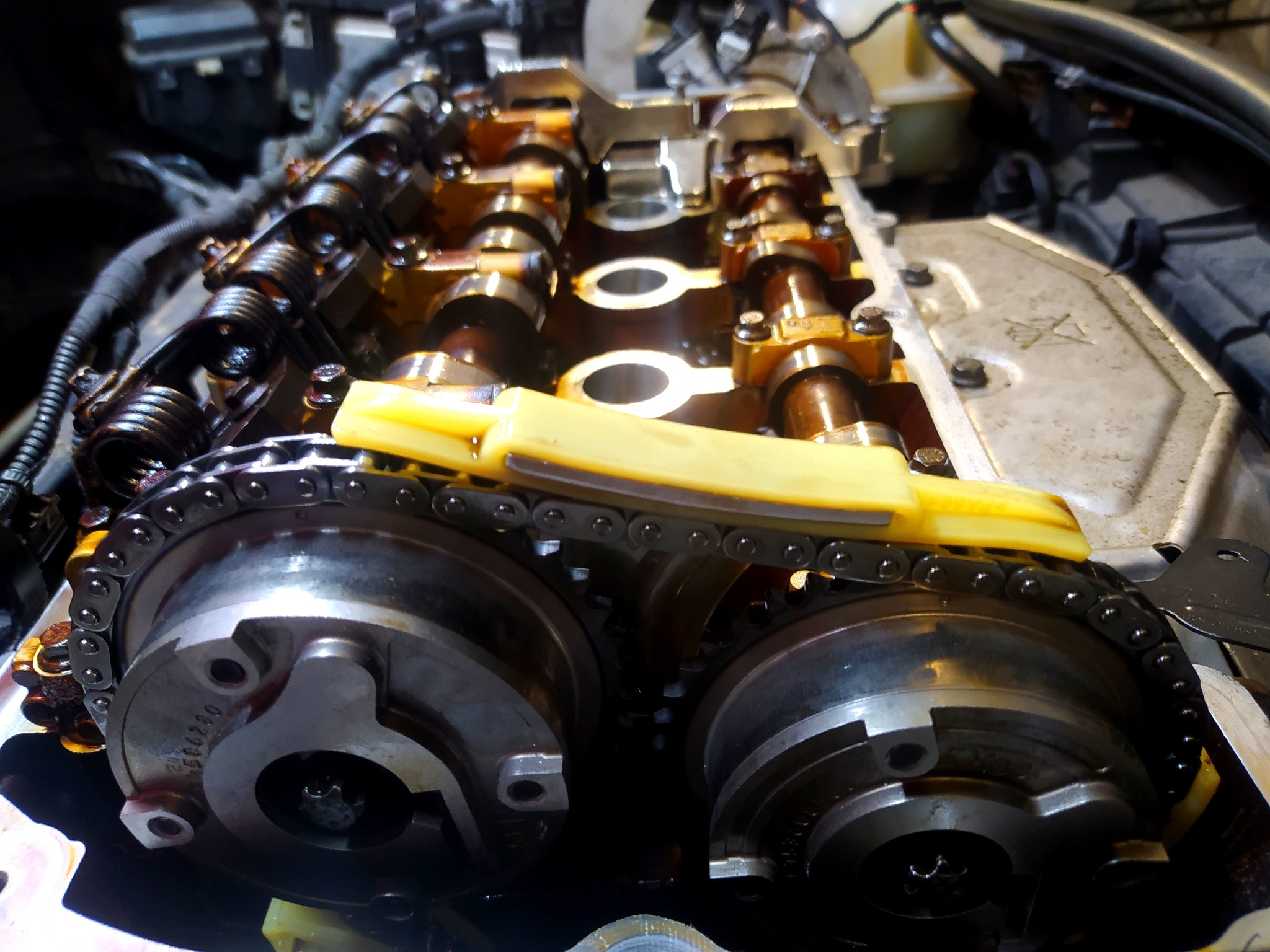 Silnik samochodowy z widocznymi wałkami rozrządu, łańcuchem rozrządu i żółtym ślizgiem, podczas naprawy lub przeglądu, oświetlenie warsztatowe.