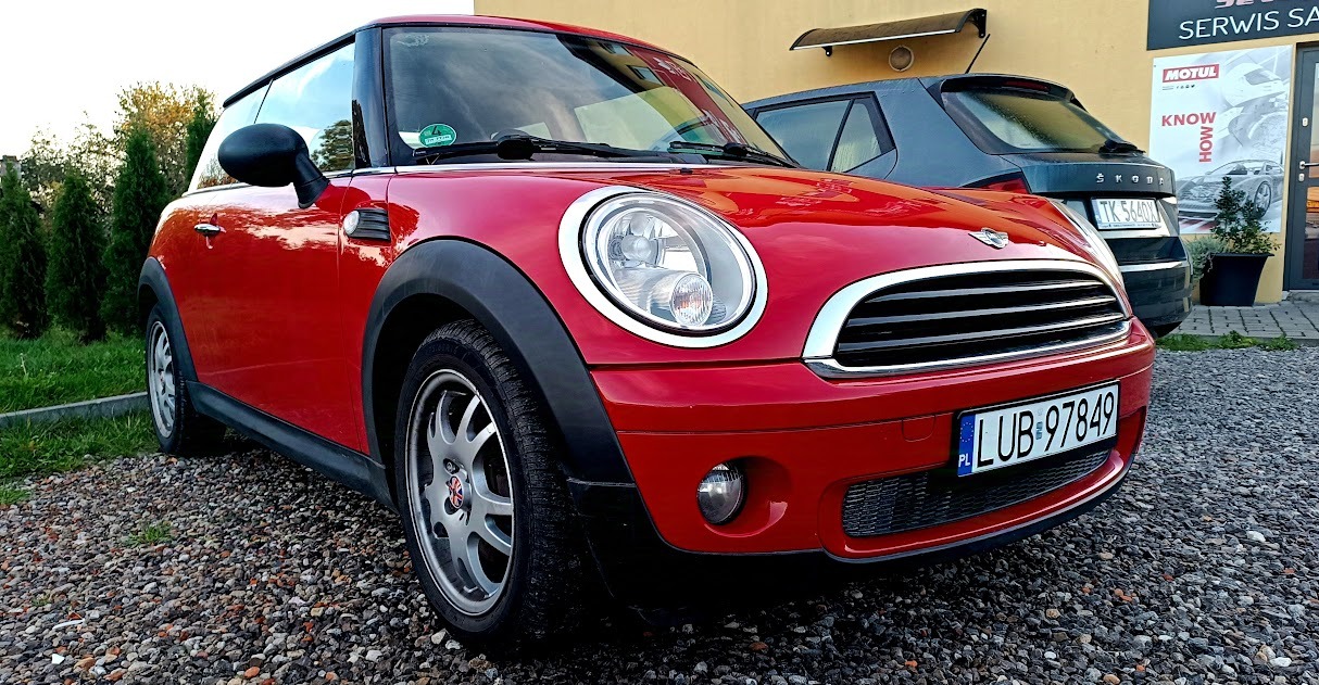 Czerwony Mini Cooper zaparkowany na żwirowym parkingu przed warsztatem samochodowym, widoczny plakat Motul i Skoda w tle.
