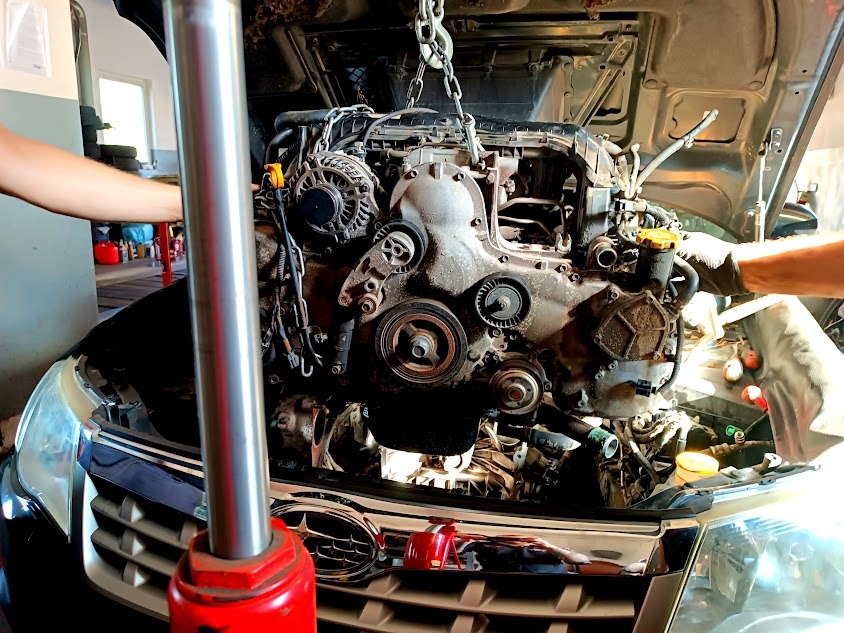 Silnik samochodowy podwieszony na łańcuchu podczas demontażu z pojazdu Subaru, widoczne ręce dwóch osób pracujących przy silniku, z przodu czerwony podnośnik hydrauliczny.