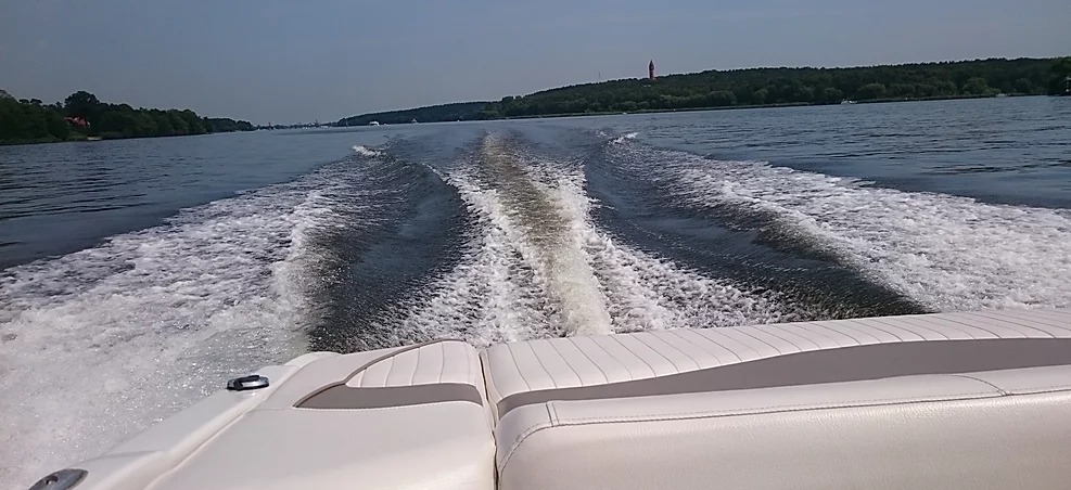 Widok z rufy motorówki na wzburzoną wodę, tworzącą białą pianę, za nią rozciąga się krajobraz zalesionych brzegów i odległej wieży.