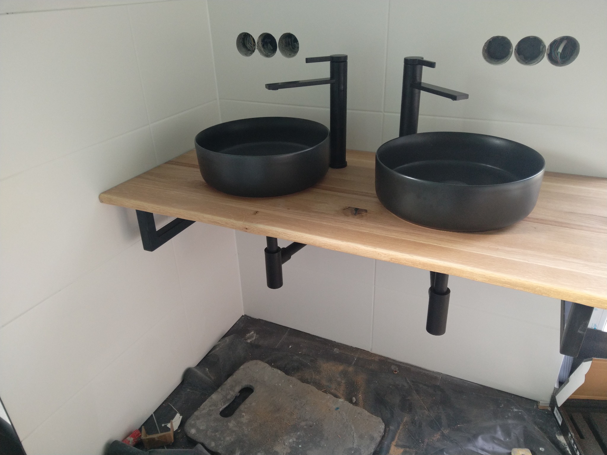 Dwie czarne umywalki nablatowe o okrągłym kształcie z czarnymi bateriami, zamontowane na drewnianym blacie w łazience w trakcie remontu, widoczne przygotowane otwory w ścianie na rury.