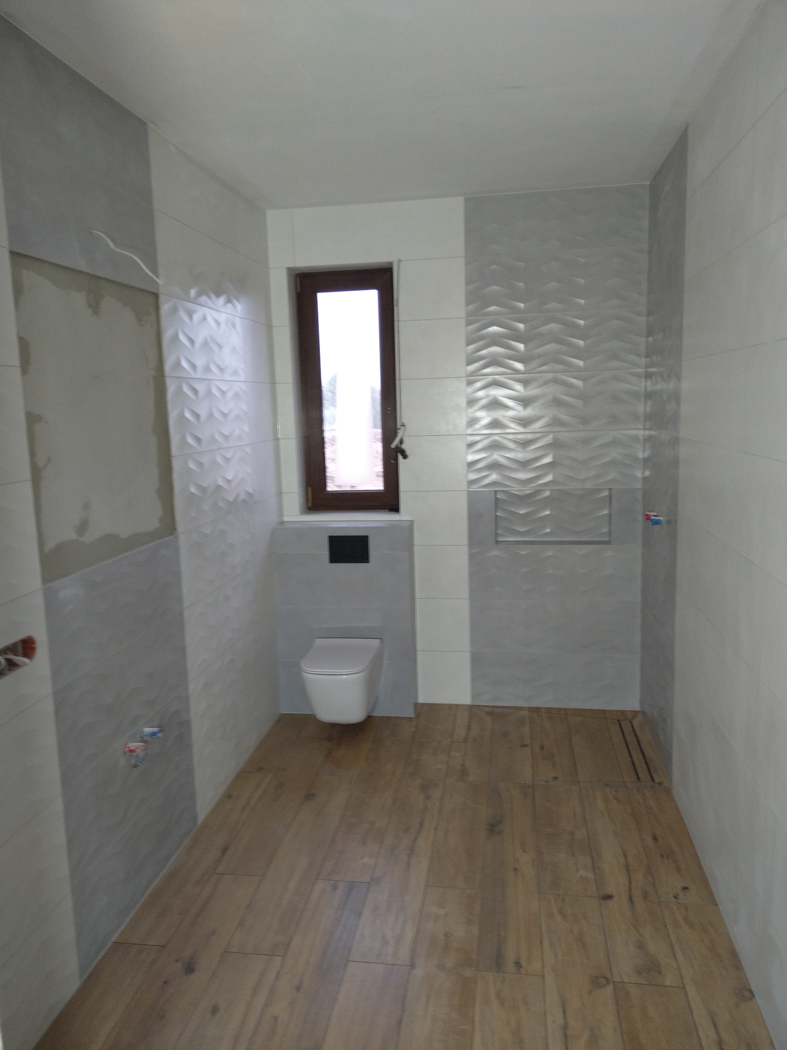 Wnętrze łazienki w trakcie wykańczania: częściowo położone szare płytki strukturalne na ścianach, drewnopodobna podłoga, wisząca toaleta, okno z brązową ramą, widoczne niedokończone fragmenty...