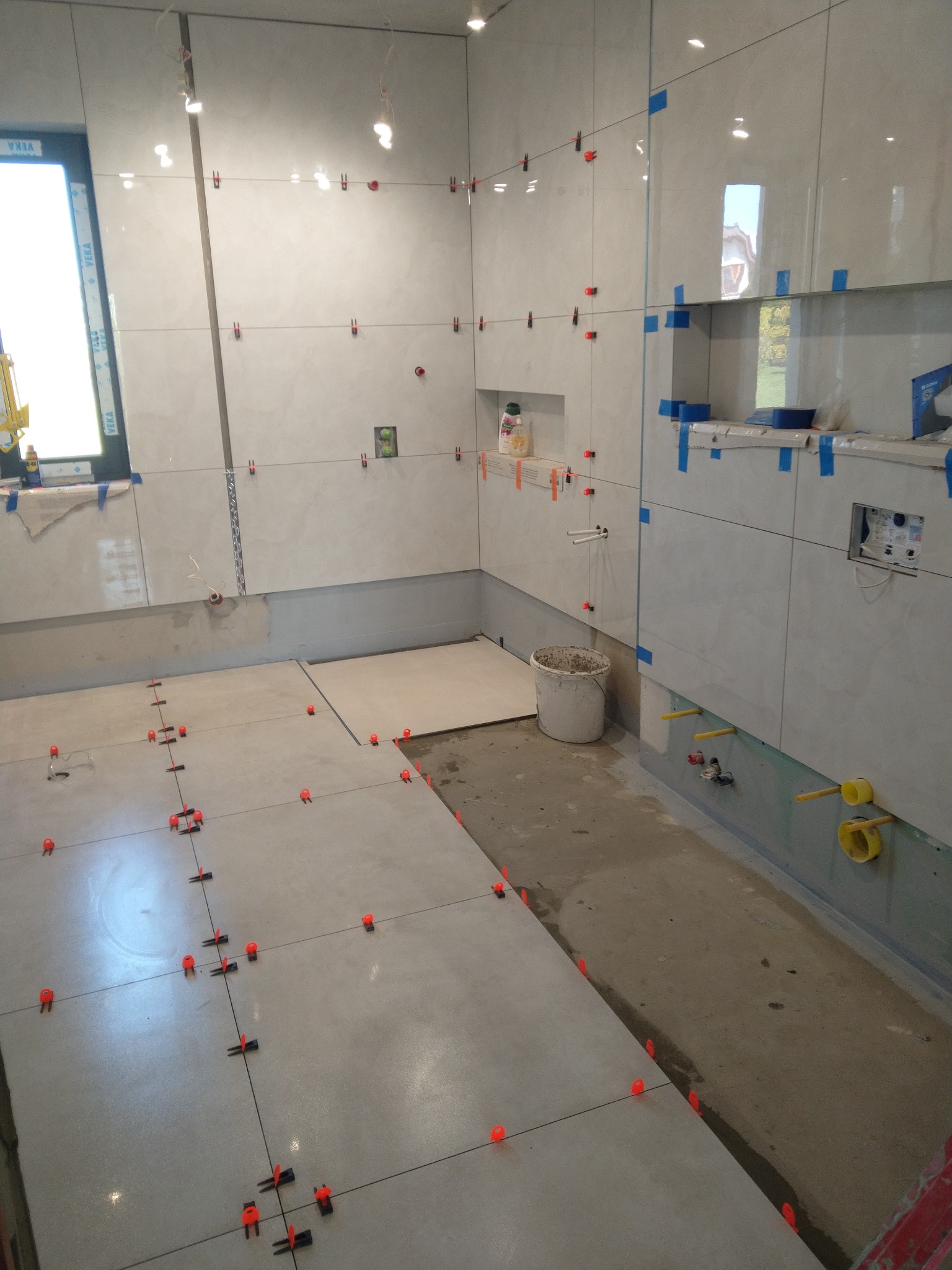 Łazienka w trakcie układania płytek podłogowych i ściennych, widoczne klipsy poziomujące, odkryte instalacje wodne i częściowo wykończona wnęka