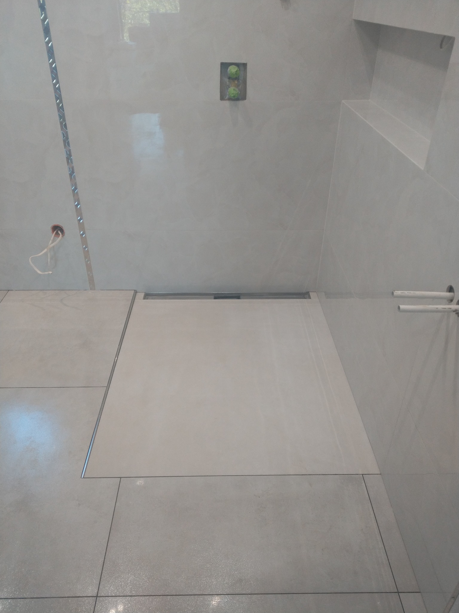 Wykończona strefa prysznicowa z odpływem liniowym, jasne płytki ceramiczne na ścianach i podłodze, widoczne przyłącza wodne i instalacja elektryczna w trakcie prac wykończeniowych.