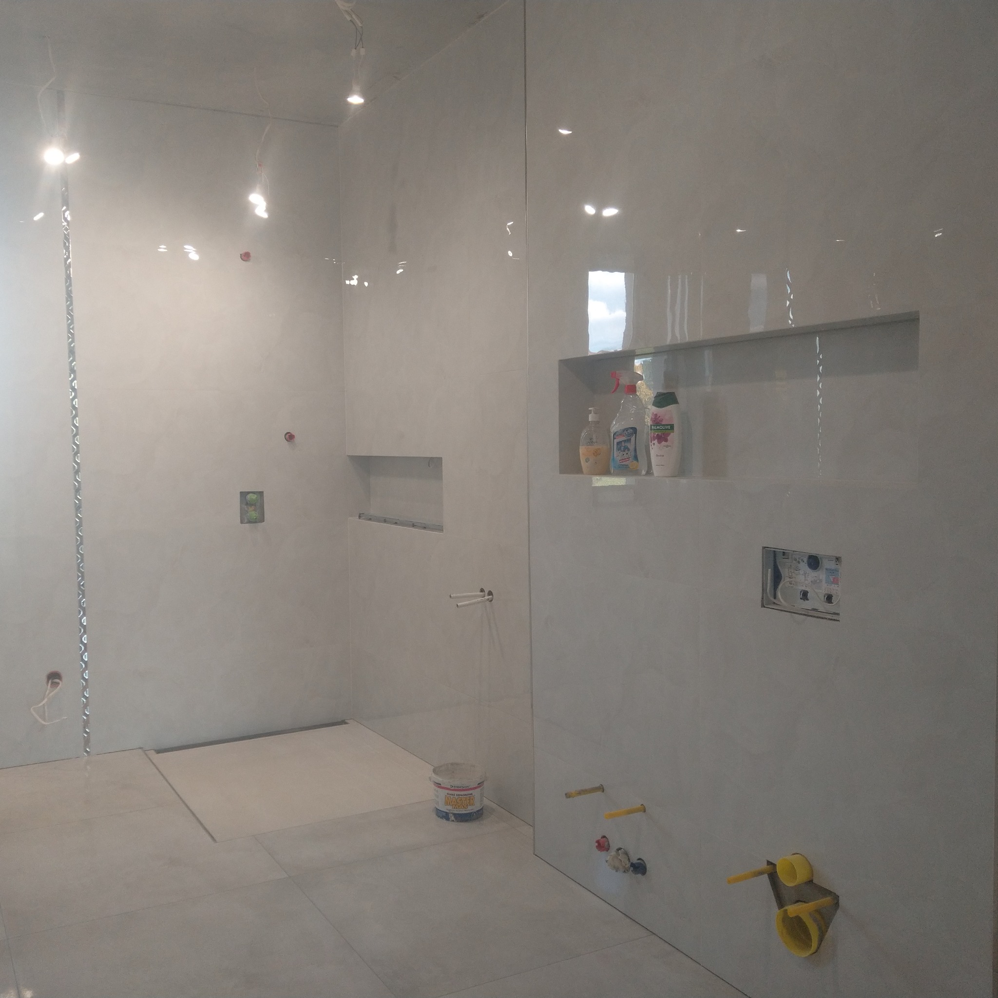 Łazienka w trakcie wykańczania, widoczne białe płytki, wnęki w ścianie, przygotowane odpływy i rury, wiaderko z klejem do płytek.