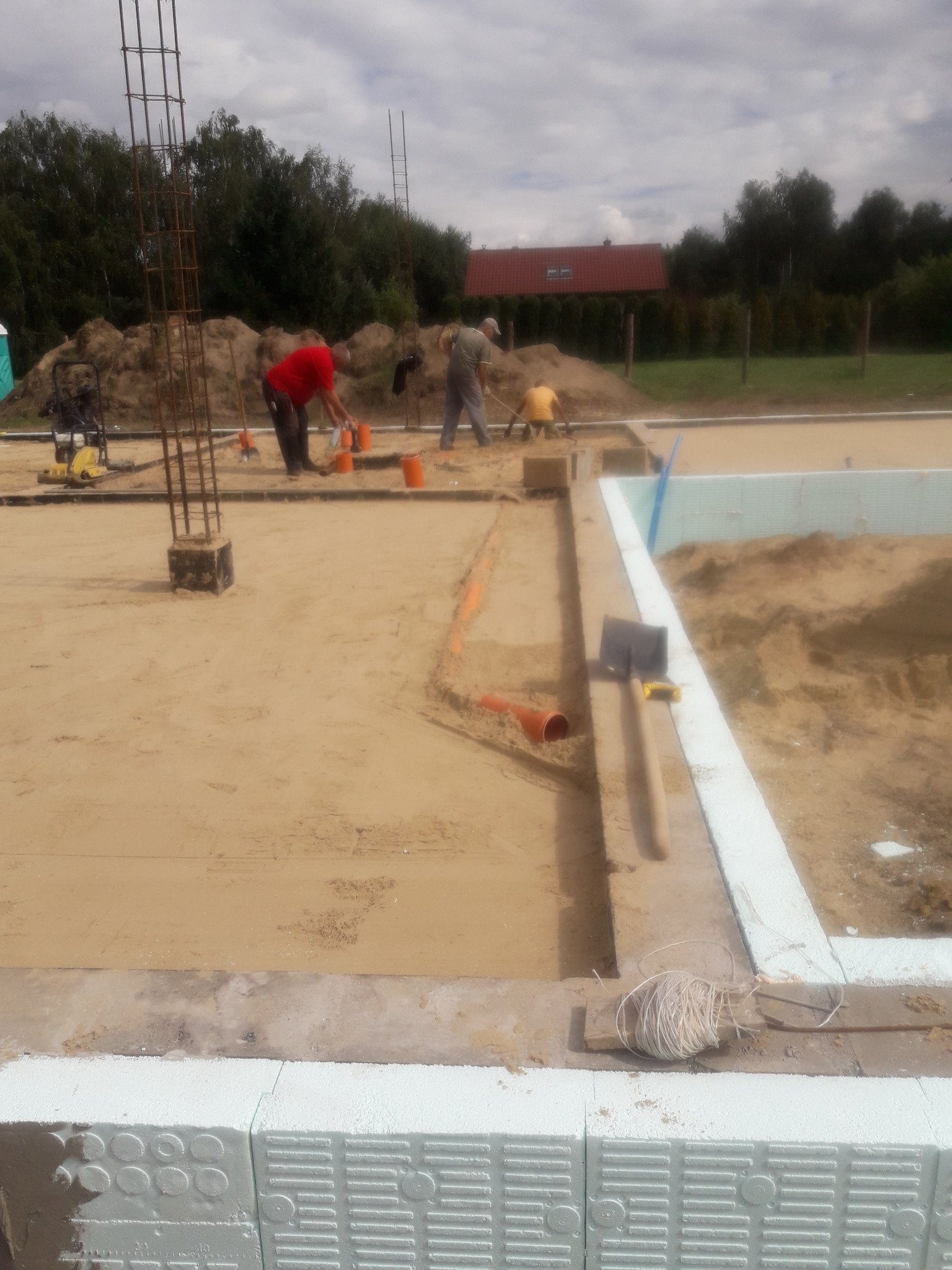 Budowa fundamentów domu: robotnicy pracują przy wylewaniu betonu, widoczne zbrojenia, szalunki z białego styropianu, rury kanalizacyjne i narzędzia budowlane.