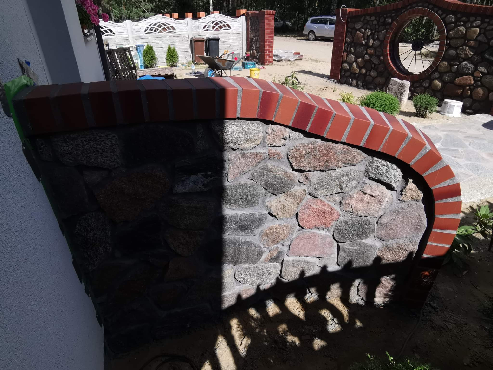 Kamienno-ceglana konstrukcja ogrodowa w półcieniu, z widocznym fragmentem białej ściany budynku i elementami ogrodu w tle.