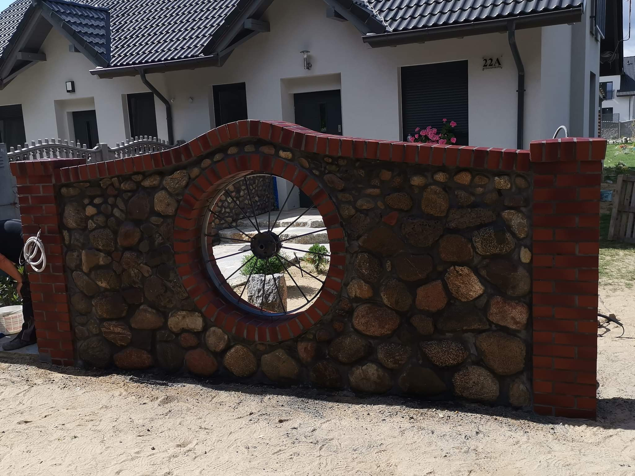 Kamienno-ceglany mur ogrodzeniowy z okrągłym oknem z metalową szprychą, na tle białego domu z ciemną dachówką.