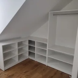 Biała szafa wnękowa z półkami i drążkiem na ubrania, dopasowana do skosu dachu na poddaszu, widok z przodu na drewnianą podłogę.