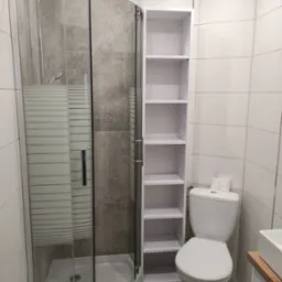 Nowoczesna łazienka z narożną kabiną prysznicową z deszczownicą, białą szafką regałową z półkami, toaletą i umywalką na drewnianym blacie, wykończona białymi i szarymi płytkami.