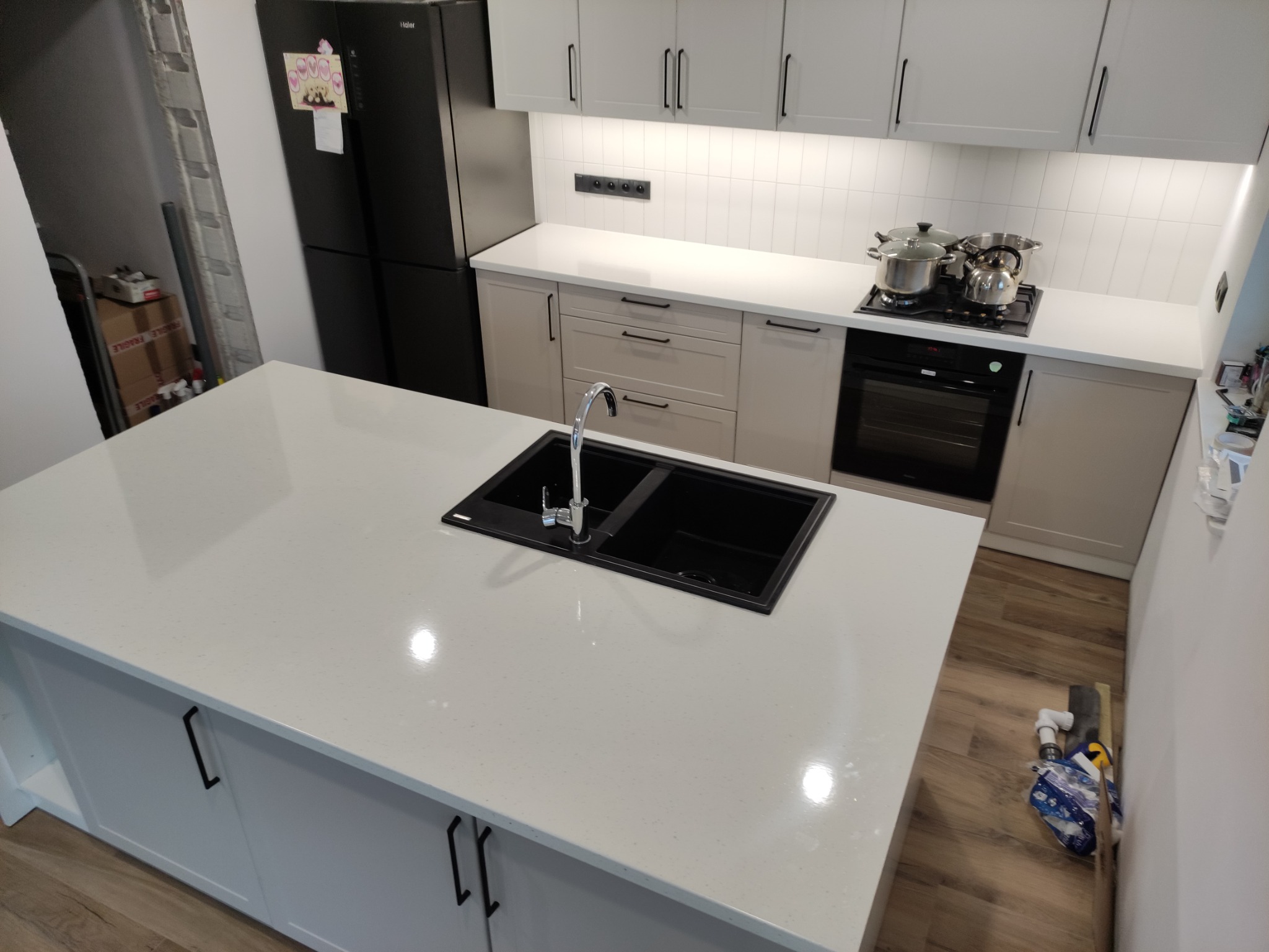 Nowoczesna kuchnia z wyspą, jasnymi szafkami z czarnymi uchwytami, czarnym zlewem i baterią, oraz czarną lodówką. Na blacie kuchennym garnki i czajnik na płycie grzewczej.