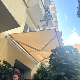 Damian Popławski - Balkon z rozłożoną, beżową markizą przeciwsłoneczną, widokiem na błękitne niebo i zieloną roślinność. Kobieta w pomarańczowej bluzce w rogu kadru.