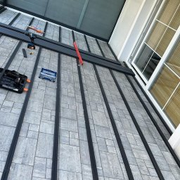 Damian Popławski - Montaż konstrukcji tarasu w Warszawie: Widok z góry na szare płyty i czarne profile, narzędzia pracy widoczne w kadrze, okno w tle.