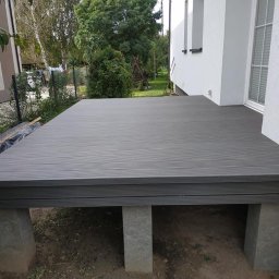 Damian Popławski - Szary taras na betonowych podporach przy białym budynku. Deski z wyraźnym wzorem drewna. Widoczne elementy ogrodu i żółta skrzynka gazowa.