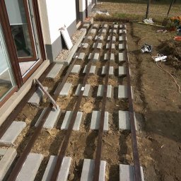 Damian Popławski - Przygotowanie podłoża pod taras: betonowe bloczki ustawione w rzędach, metalowe legary, ziemia. Widoczne narzędzia i fragment budynku.