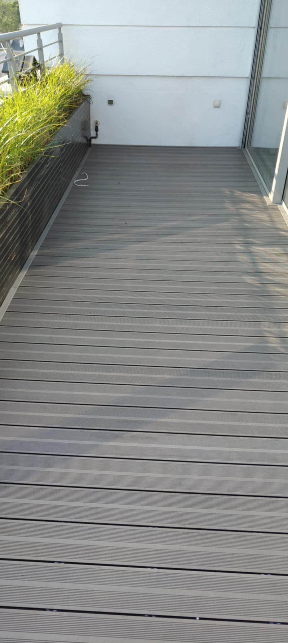 Szary taras z desek kompozytowych, z wyeksponowaną donicą z zieloną trawą i białym murem w tle, oświetlony naturalnym światłem.